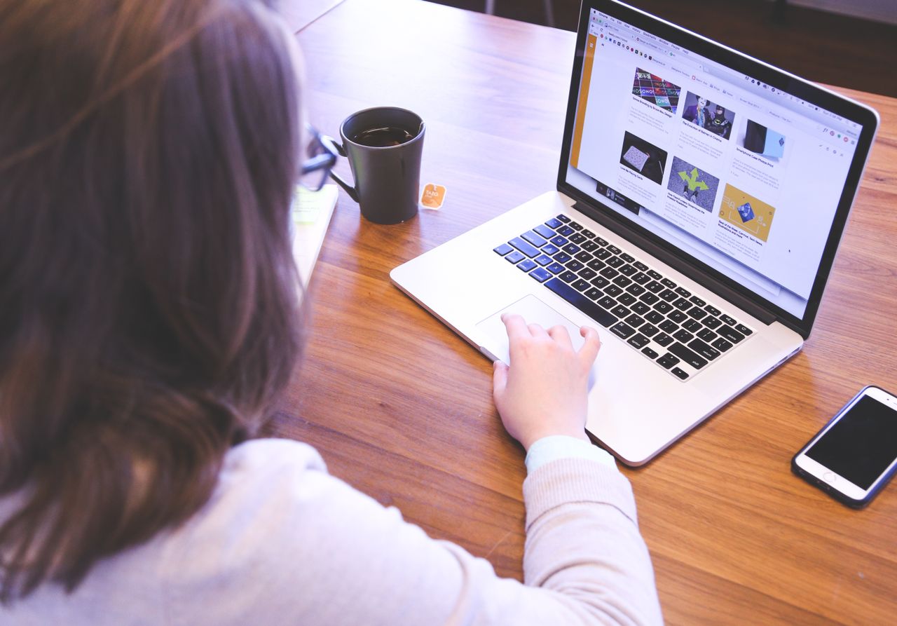 Une personne naviguant sur le web sur un MacBook posé sur un bureau en bois, avec un smartphone.